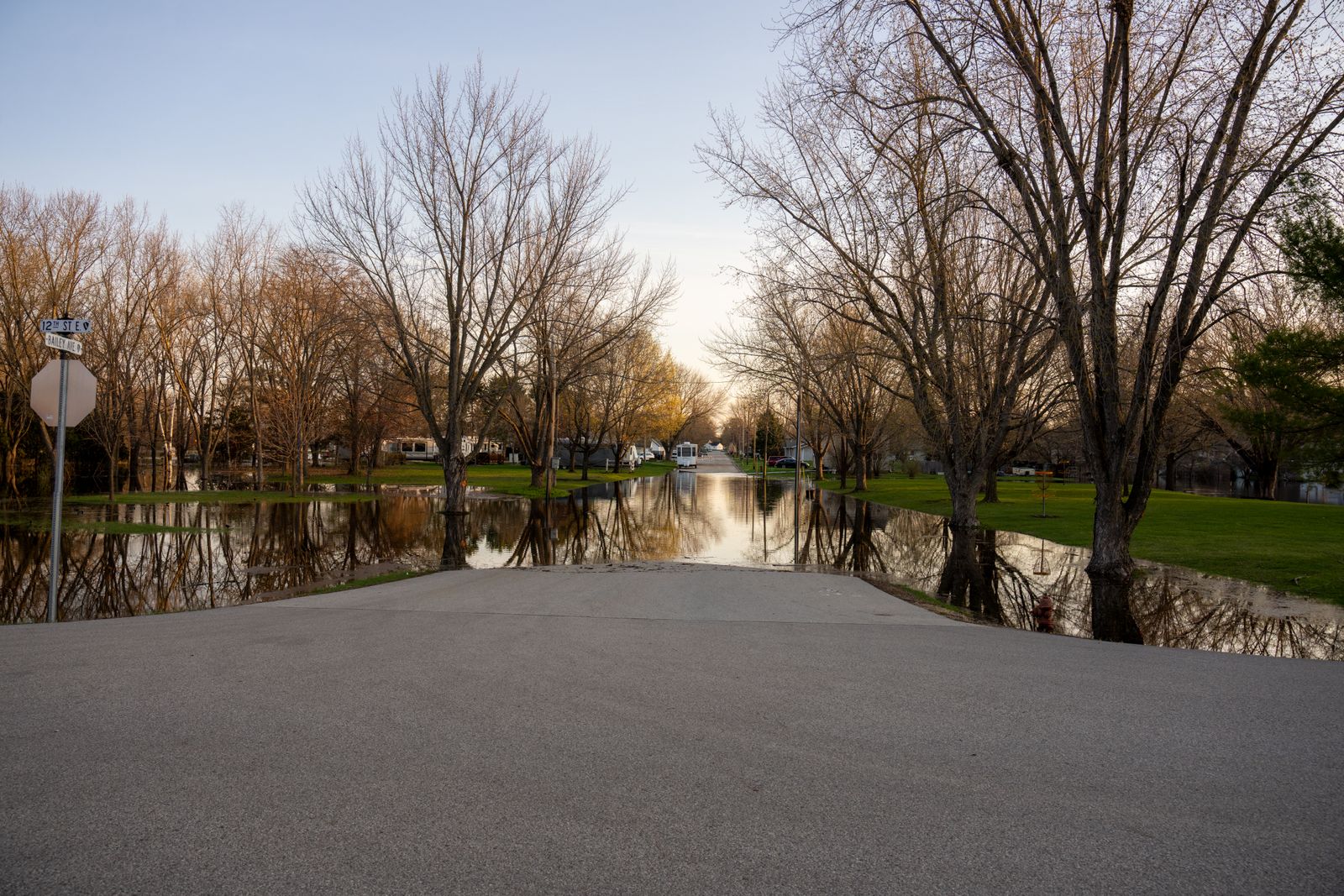 Floodwater covering a park and roadway with bare trees reflected in the water