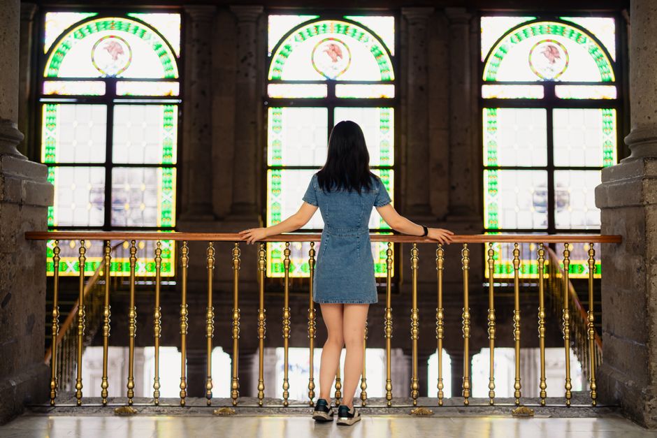 Mujer de pie junto a una barandilla ornamentada con coloridos vitrales en un palacio histórico mexicano.