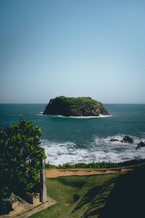 Free A picturesque view of a small rocky island surrounded by the ocean, framed by lush greenery and sea waves. Stock Photo