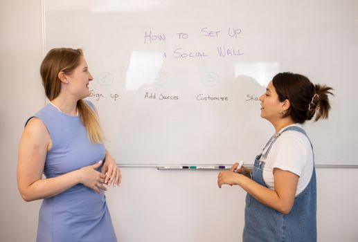Two women discussing social media wall setup with tips on a whiteboard.