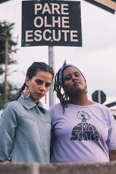 Two women pose under a street sign reading 'Pare Olhe Escute' outdoors.