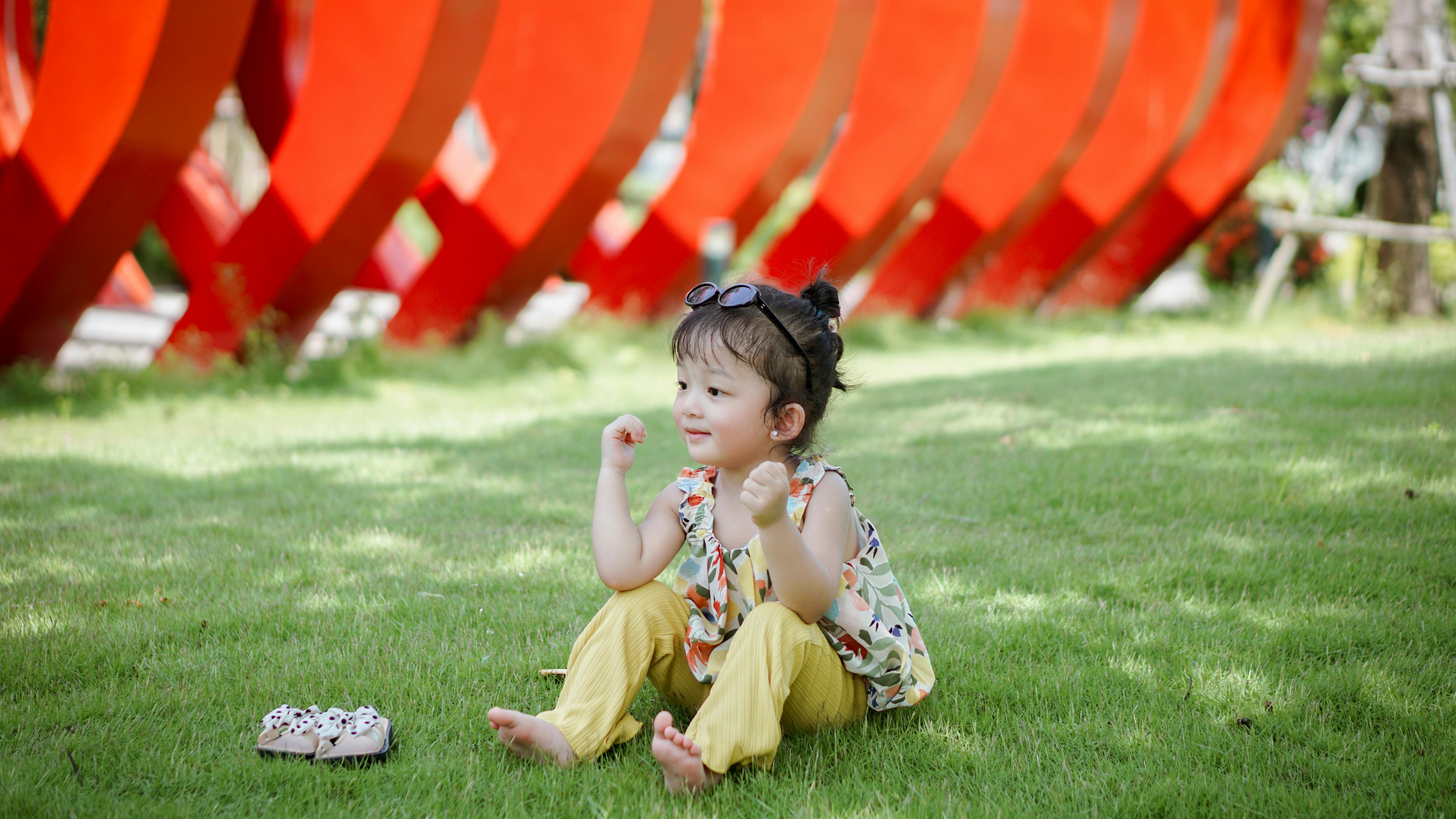 a little girl sitting on the grass in front of a red sculpture