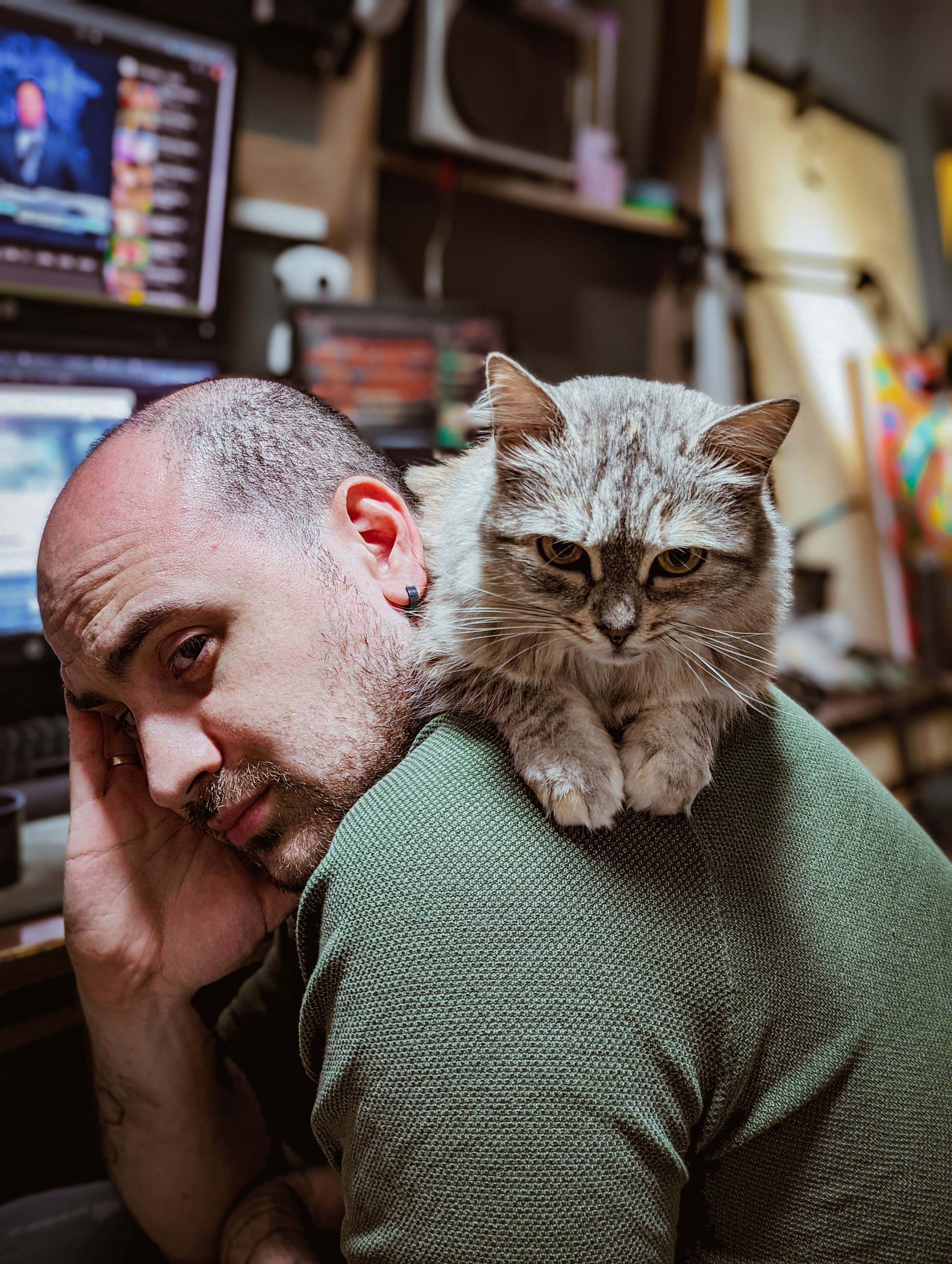 A man resting with a fluffy cat on his shoulder in a cozy indoor home office setting.