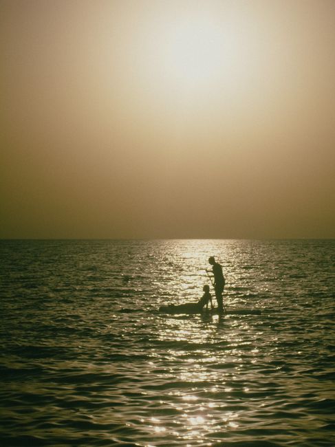 Two silhouetted people enjoy a serene sunset over a calm ocean, casting reflections on the water.