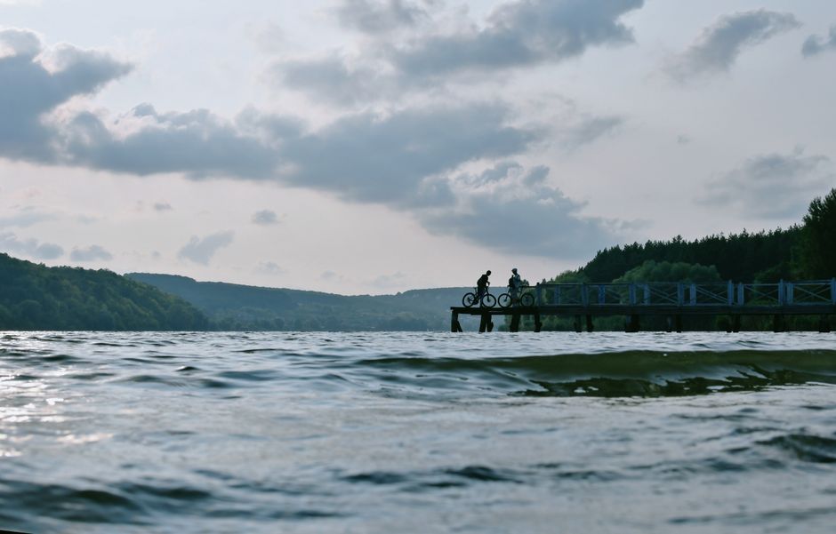 Silhouetted cyclists on a tranquil pier against a scenic lakeside backdrop.