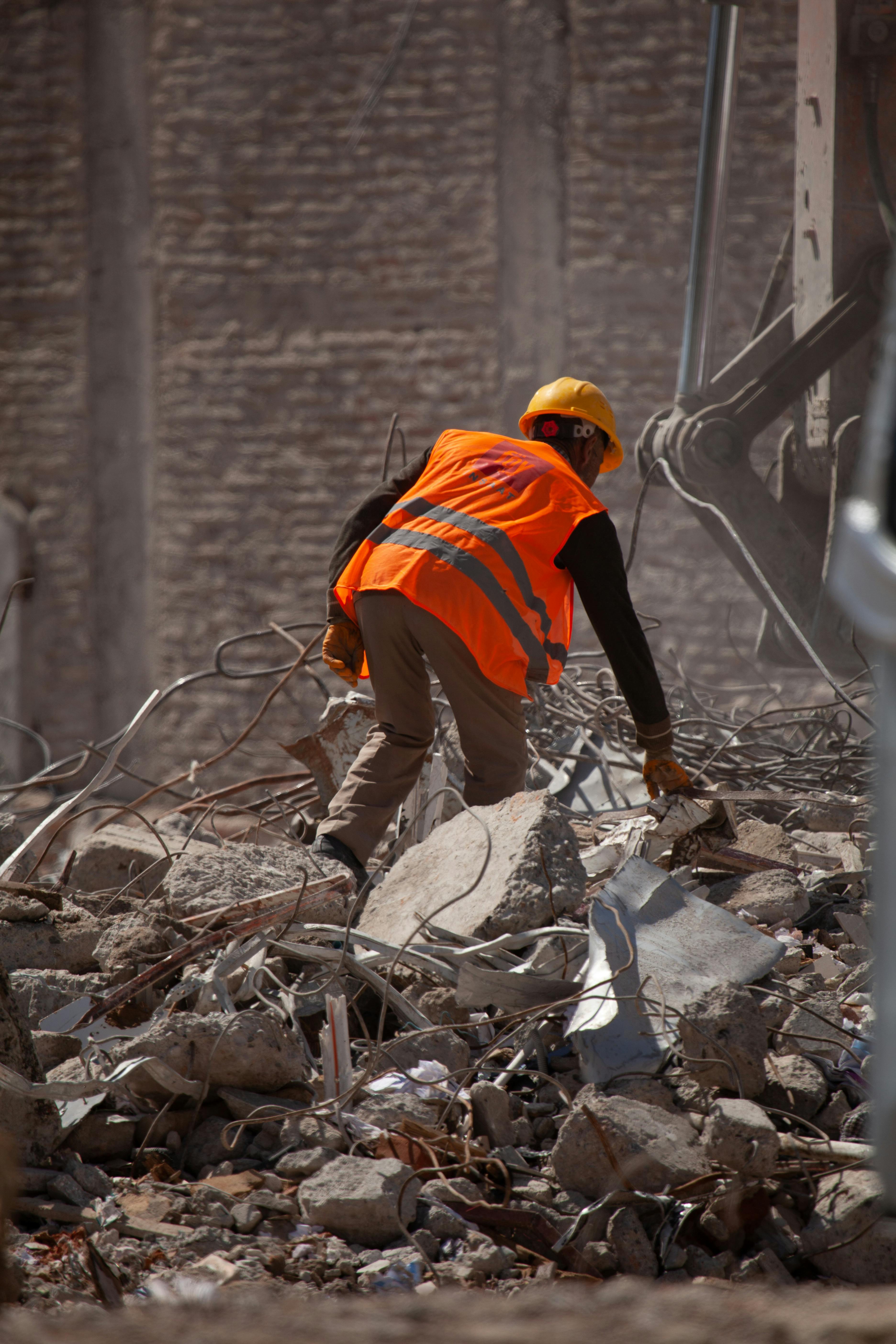 Construction worker in orange vest and hard hat searches through rubble at a building site.