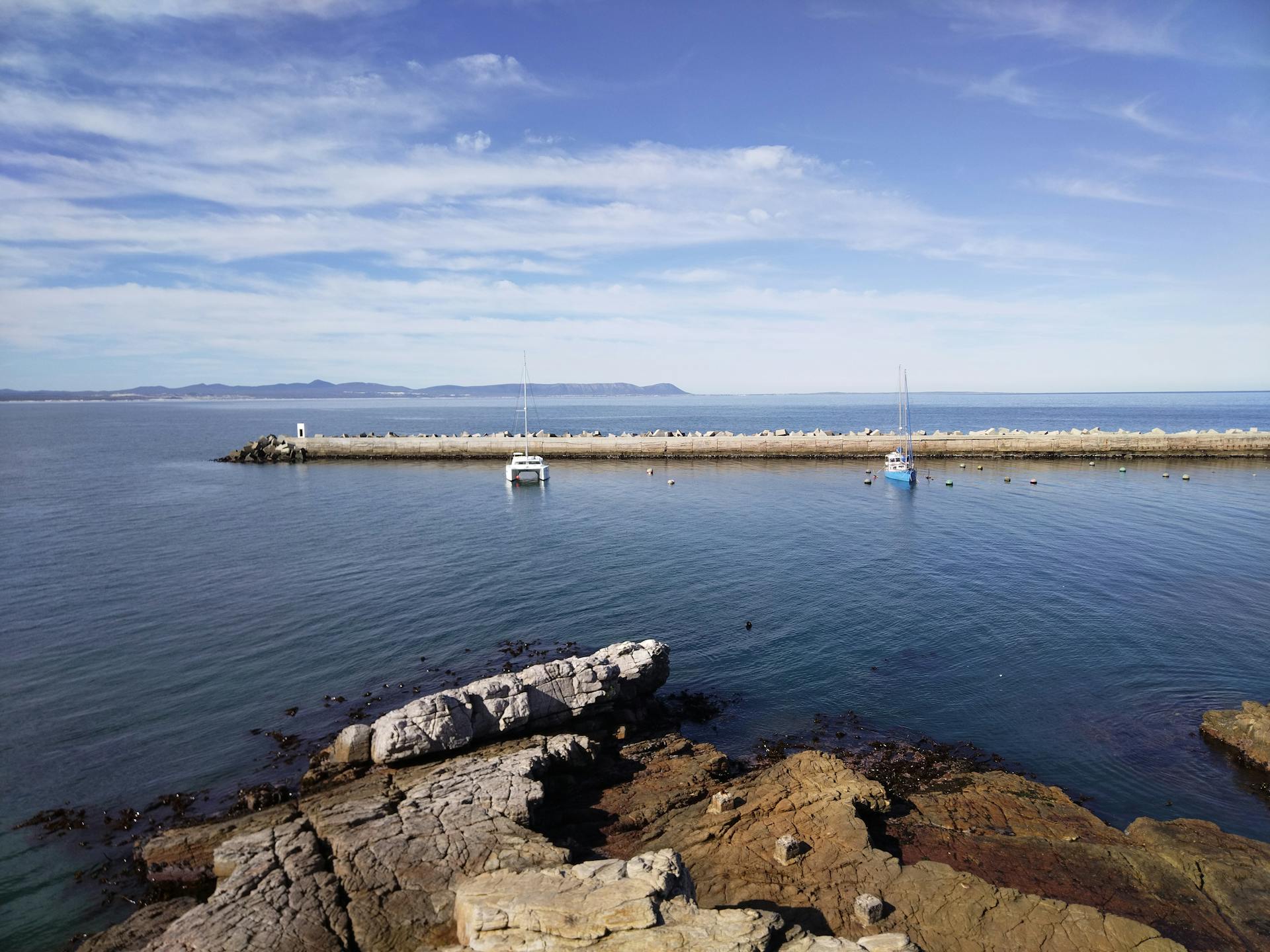 A tranquil coastal scene with rocky shoreline and boats docked by the pier under a clear blue sky.