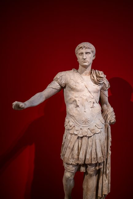 A detailed marble statue of a Roman figure against a red museum backdrop.