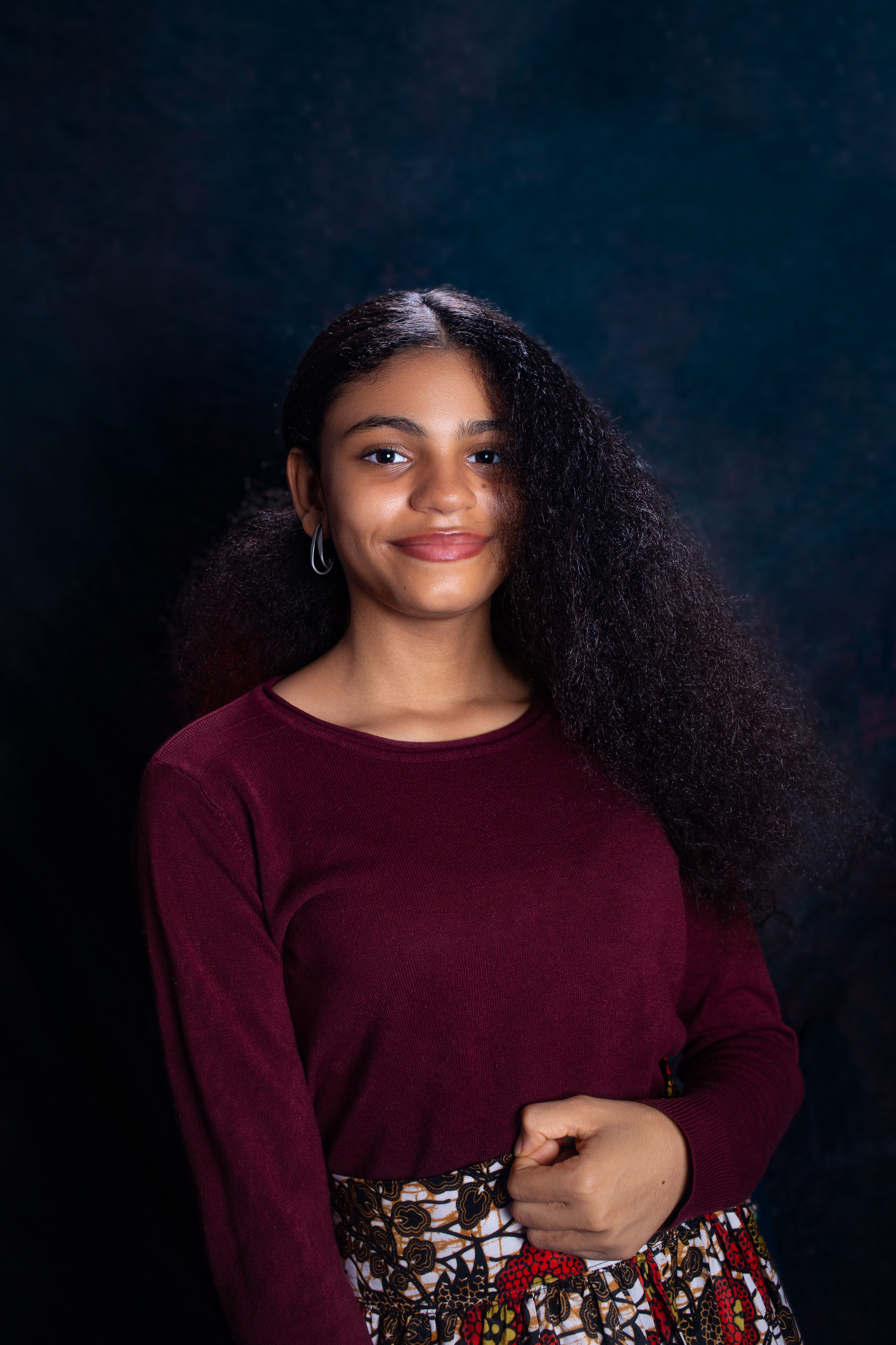 Portrait of a young woman with curly hair wearing a maroon top and a patterned skirt.