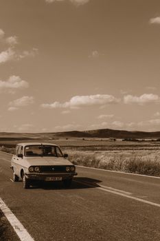 Sepia photo of a classic car driving on an empty rural road with scenic views.