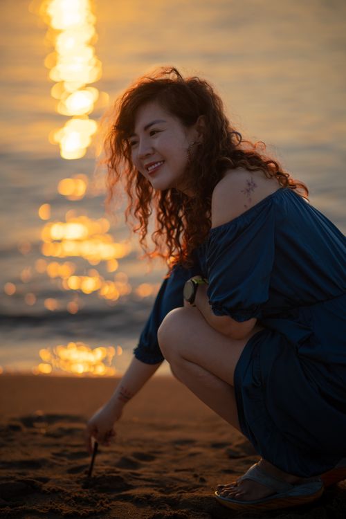 Free A smiling woman kneeling on the beach during a beautiful sunset, capturing a serene vacation moment. Stock Photo