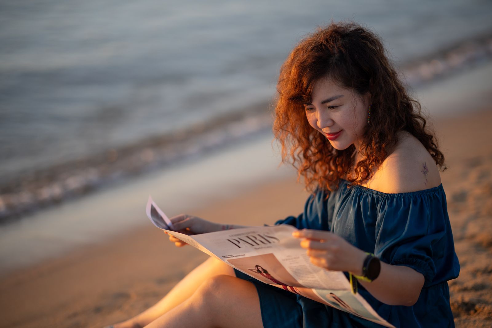Curly-haired woman in off-shoulder blue dress reads a newspaper on the beach at golden hour