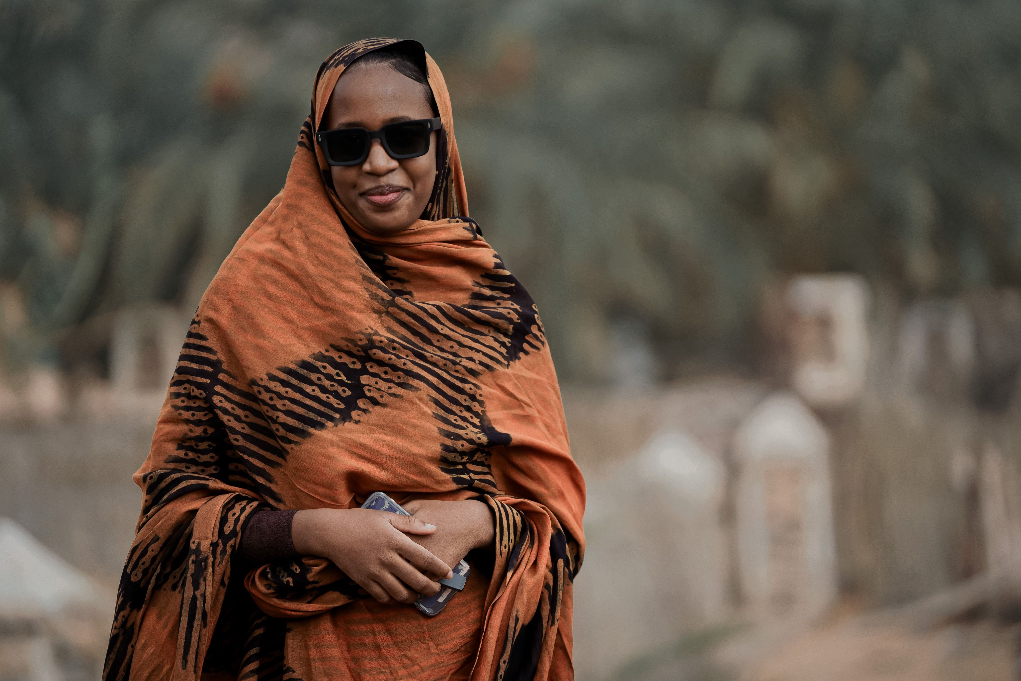 A woman wearing a traditional headscarf and sunglasses smiles confidently outdoors.