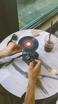 Hands handle a vintage camera with a vinyl record on a marble table by a cafe window.
