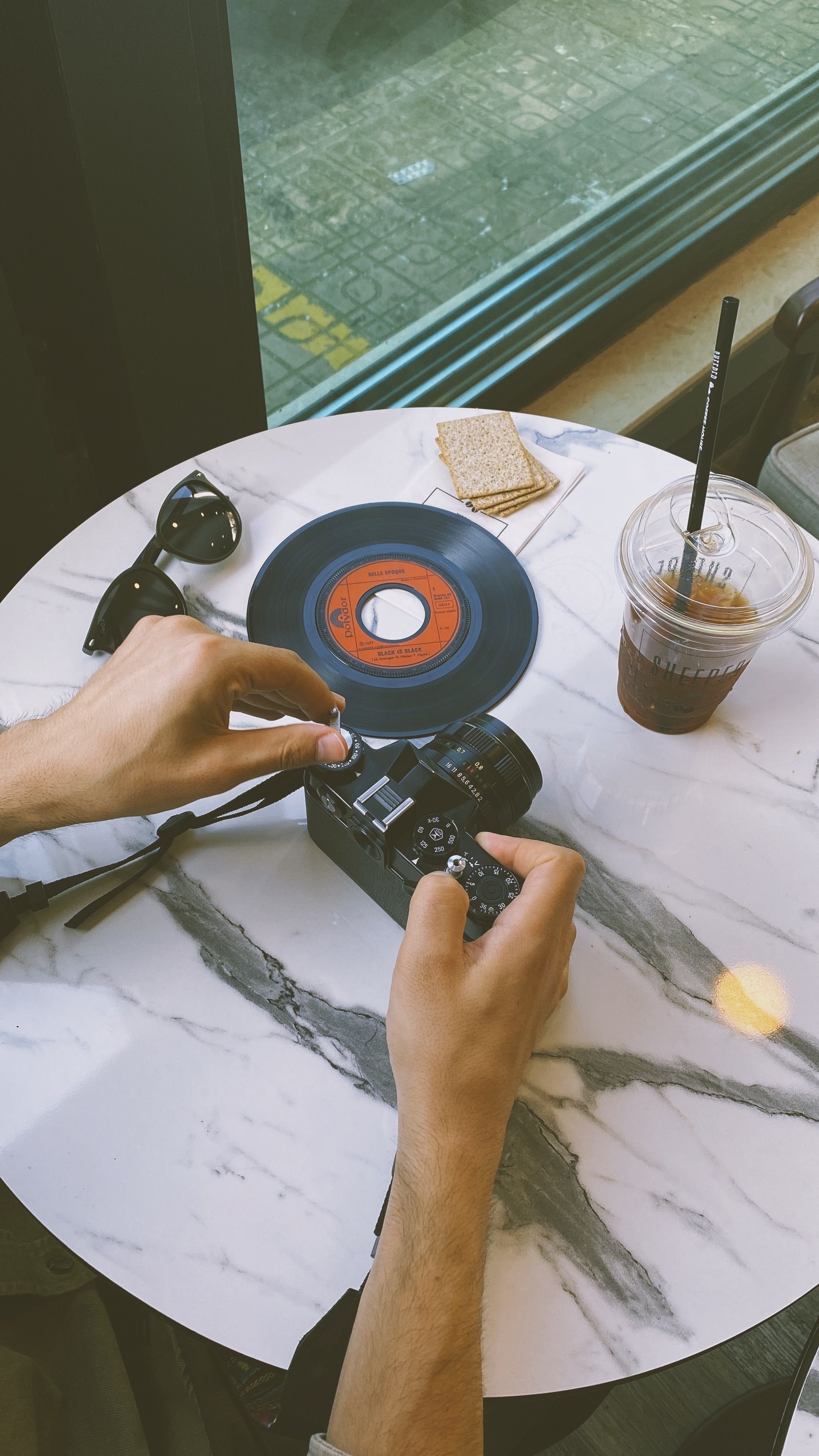 Hands holding a vintage camera on a marble table with a vinyl record and iced coffee.