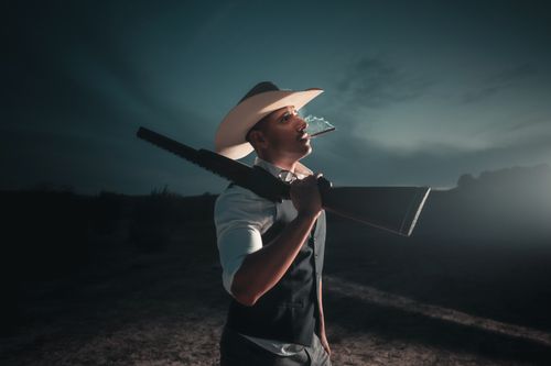 Free A cowboy with a shotgun stands smoking under a moody night sky in rural Texas. Stock Photo