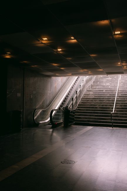 Dimly lit subway station exit featuring an escalator and stairs leading to daylight.