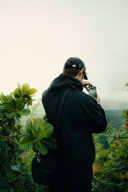 A man in a black hoodie takes photos of nature with a smartphone in an outdoor setting.
