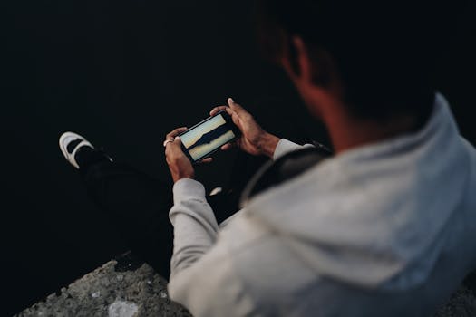 A man in casual attire sitting by the water using a smartphone, captured in a low-light setting.