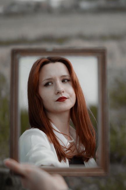 Red-haired woman holding a mirror outdoors, capturing her reflection in a serene setting.