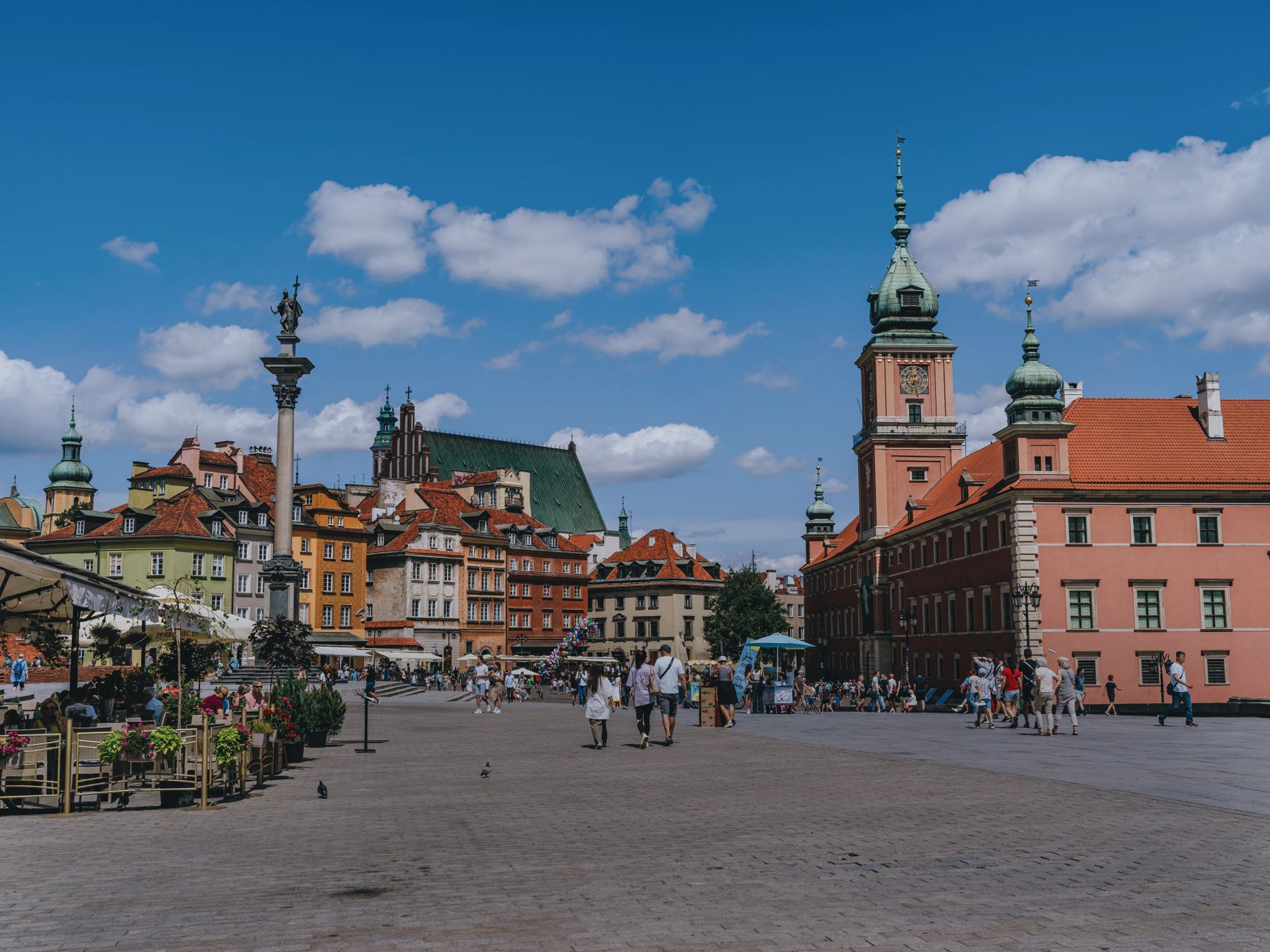 Vibrant scene of Warsaw's Old Town Square with tourists and iconic landmarks under a blue sky.