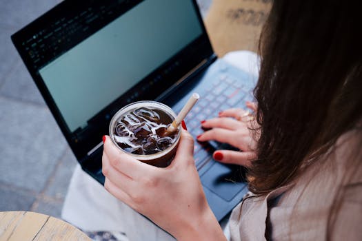 An adult woman working on a laptop while holding an iced drink, seated outside.