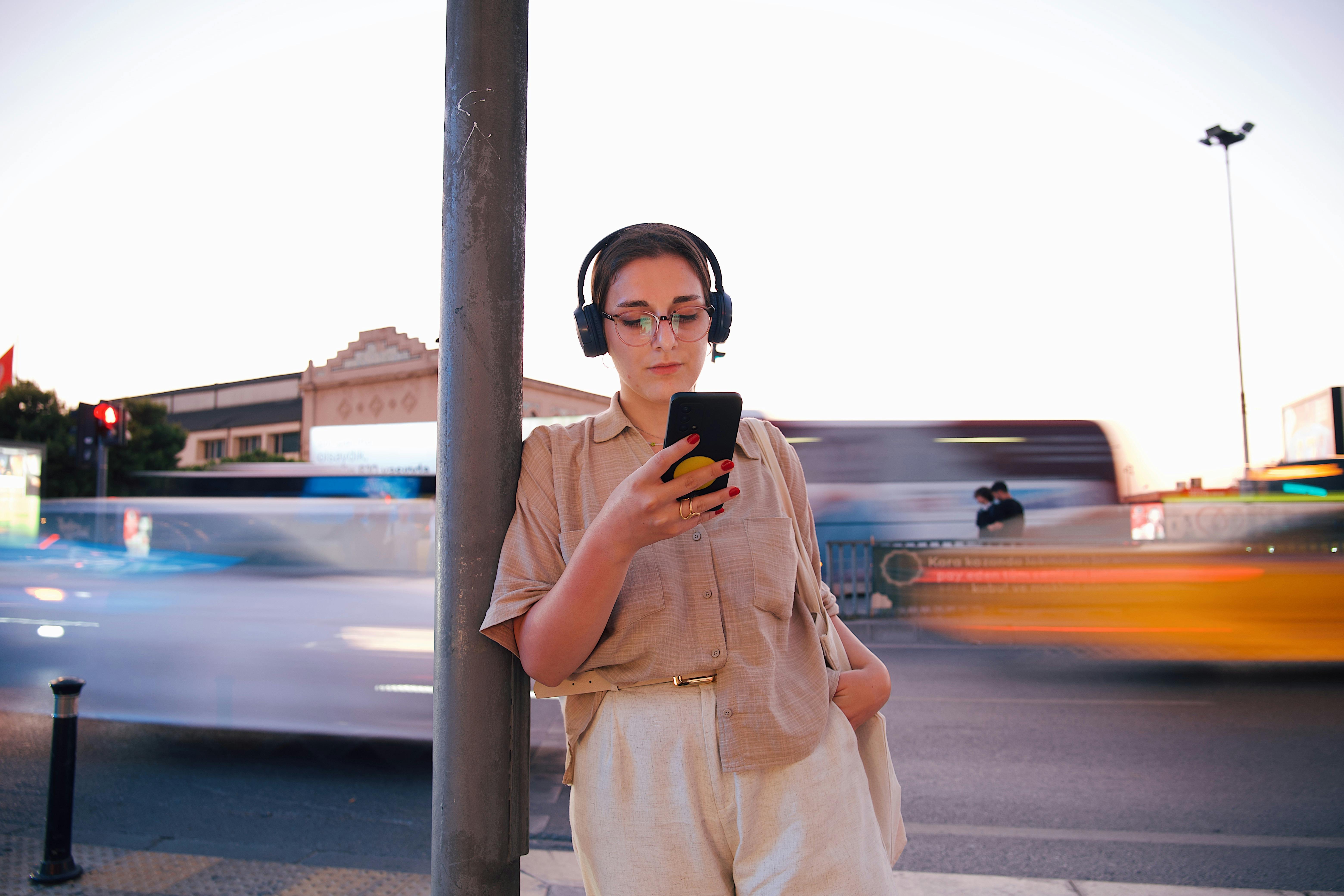 Young woman in urban setting using smartphone with headphones, traffic rushing past.