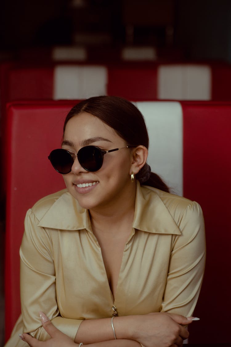 Young Woman In Sunglasses Sitting Inside And Smiling 