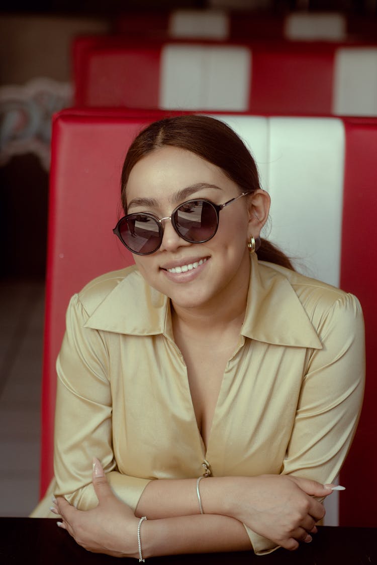 Young Woman In Sunglasses Sitting Inside And Smiling 