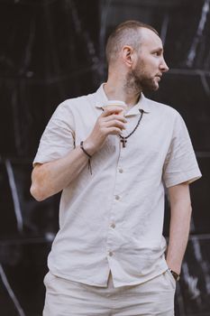 Casual urban portrait of a man in white attire, holding coffee, looking away.