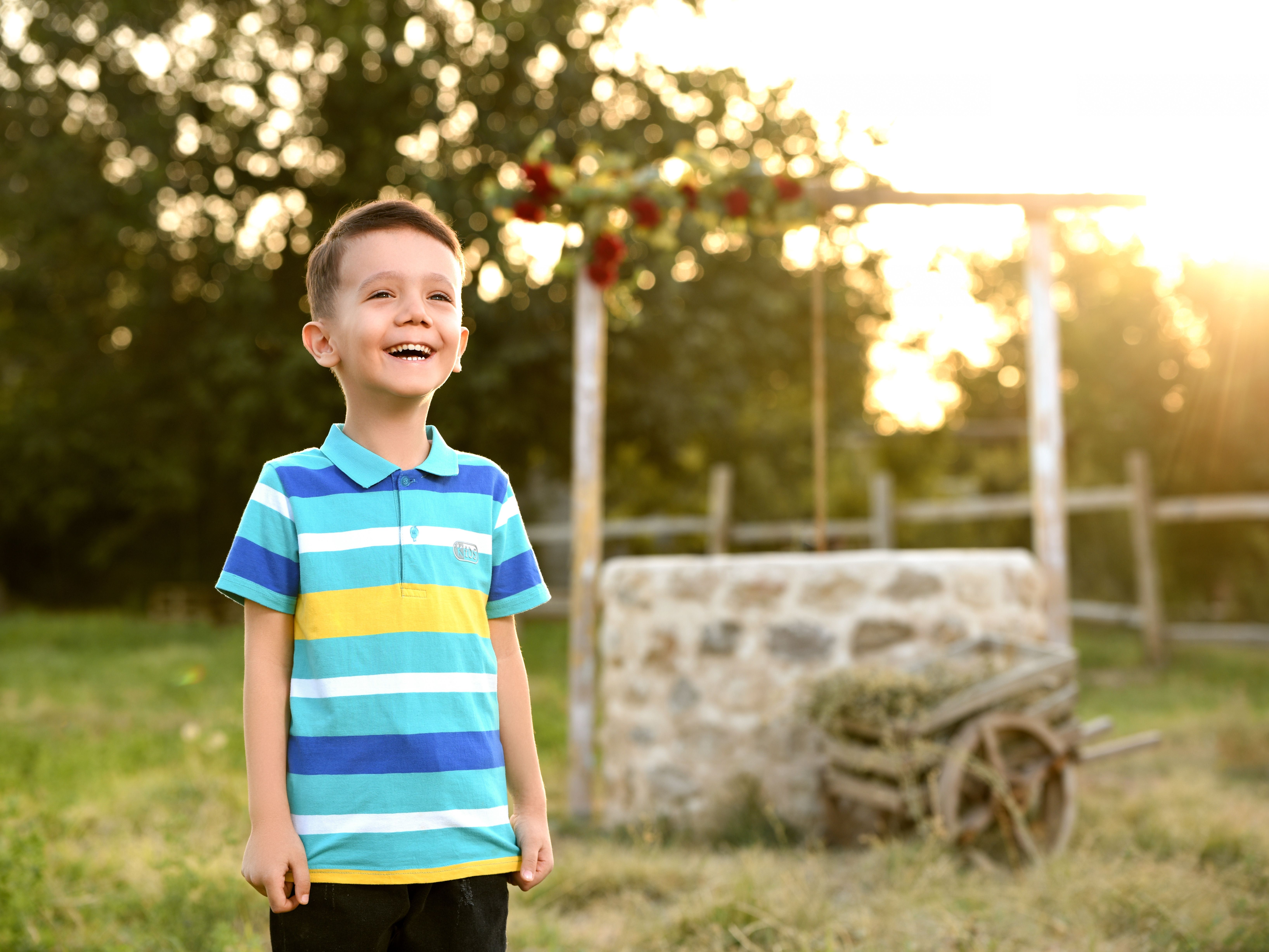 A cheerful boy in a striped shirt smiles joyfully in a sunny garden setting during sunset.