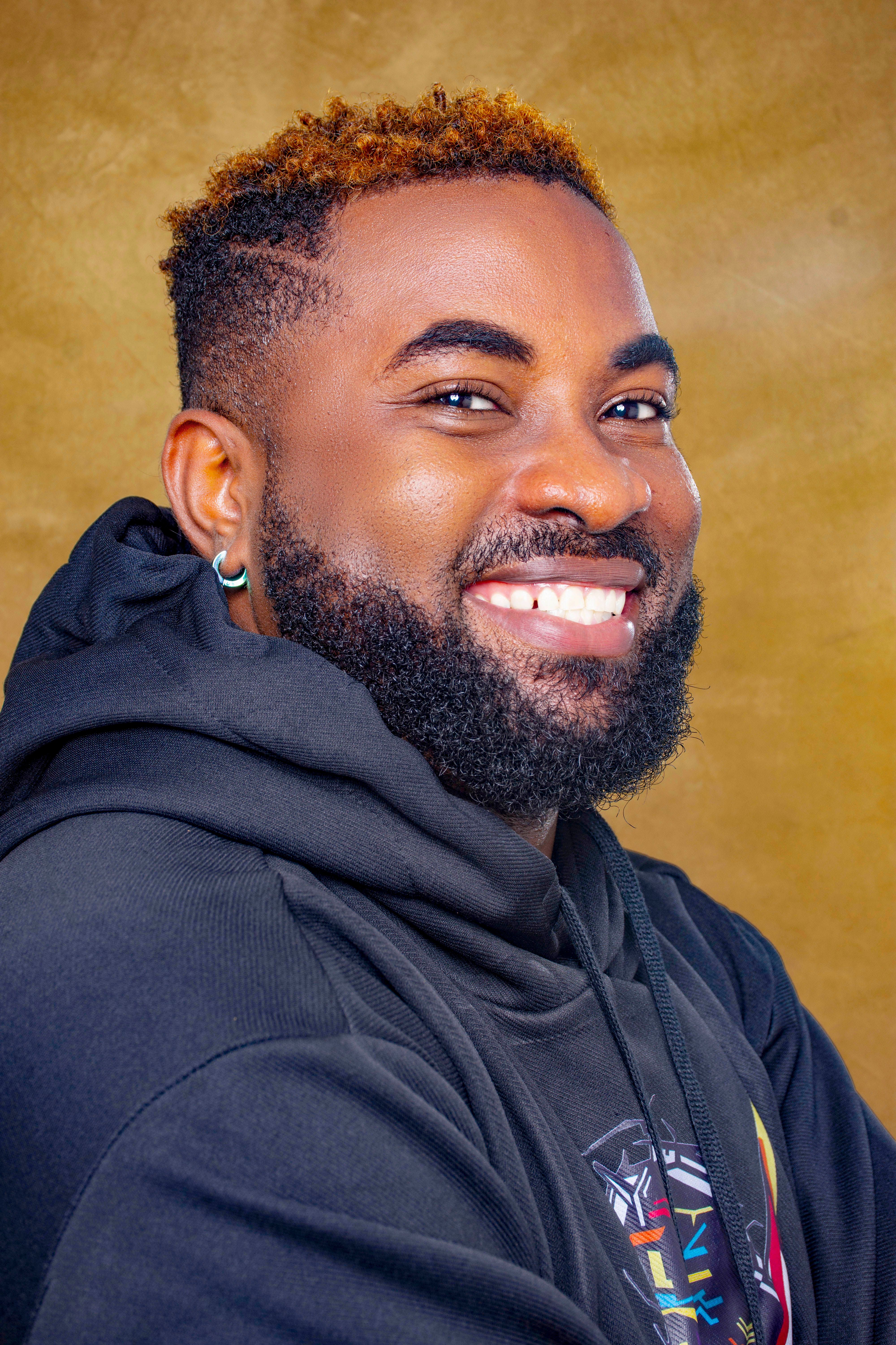 A cheerful young man with a beard wearing a stylish hoodie, smiling against a studio background.