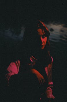 Dramatic nighttime portrait of a young woman sitting by a river with a moody atmosphere.