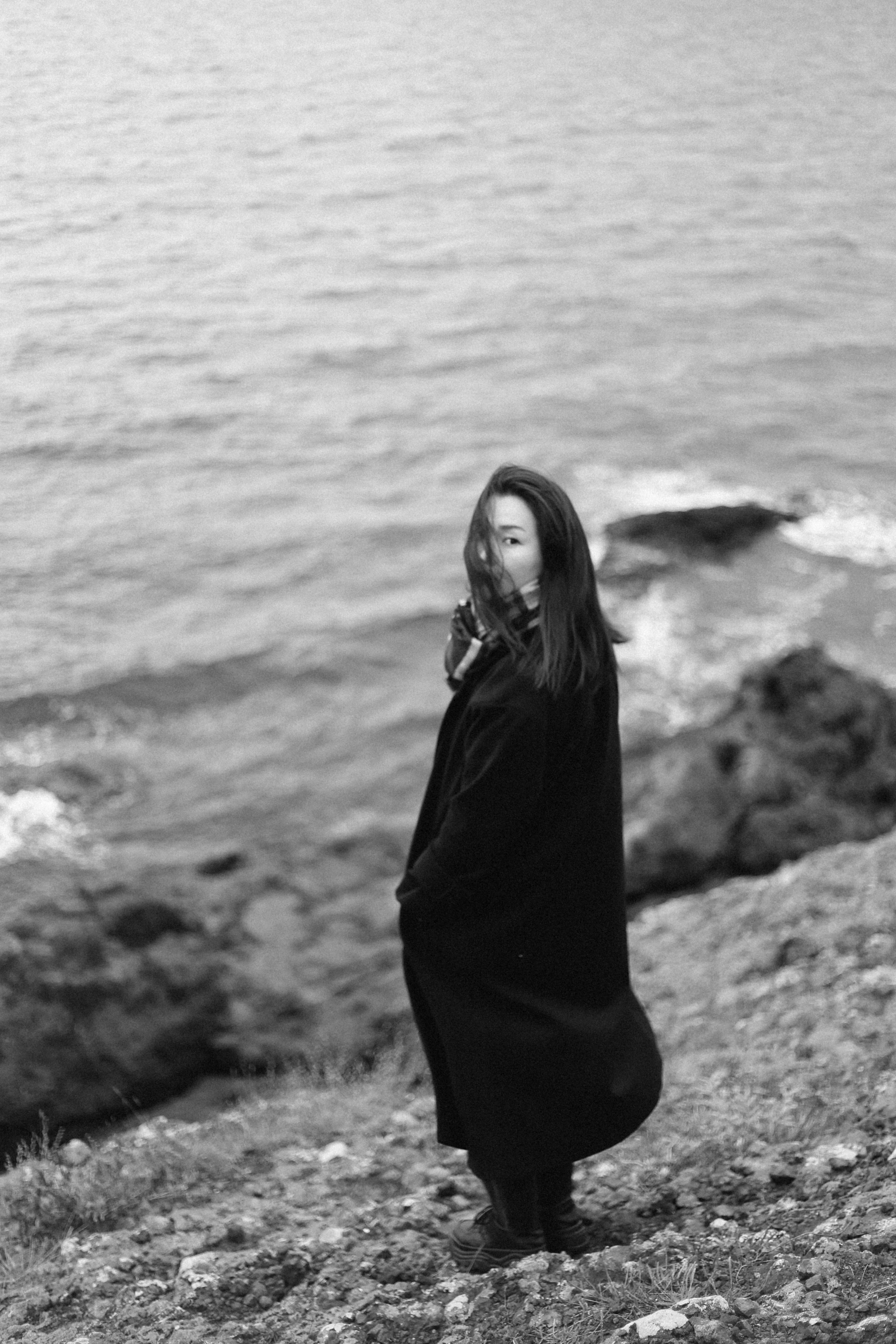 A woman in a coat stands on a rocky shore, gazing back towards the camera.