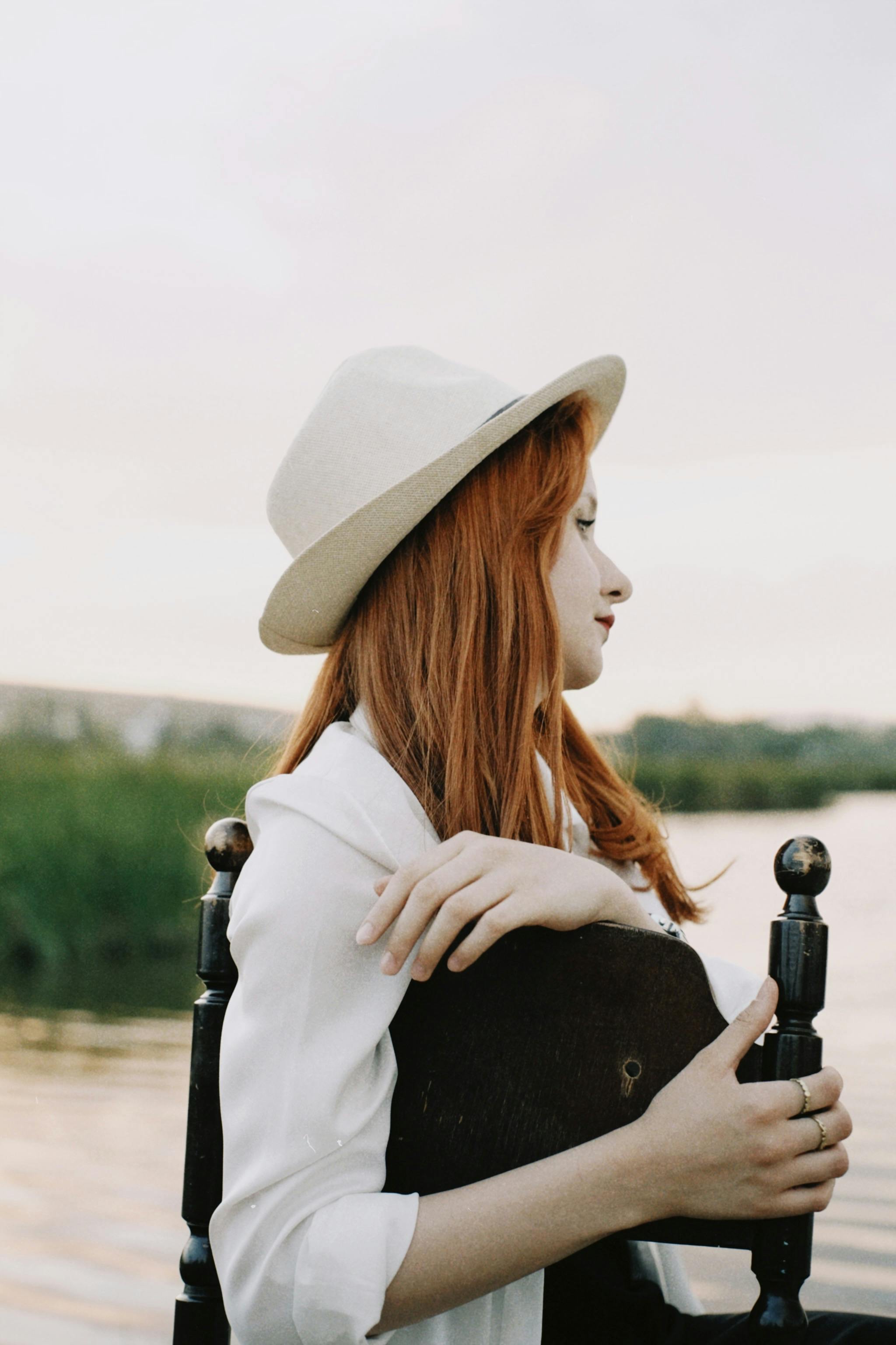 Side profile of a redhead woman in a hat, sitting by a tranquil lake, creating a serene and contemplative mood.