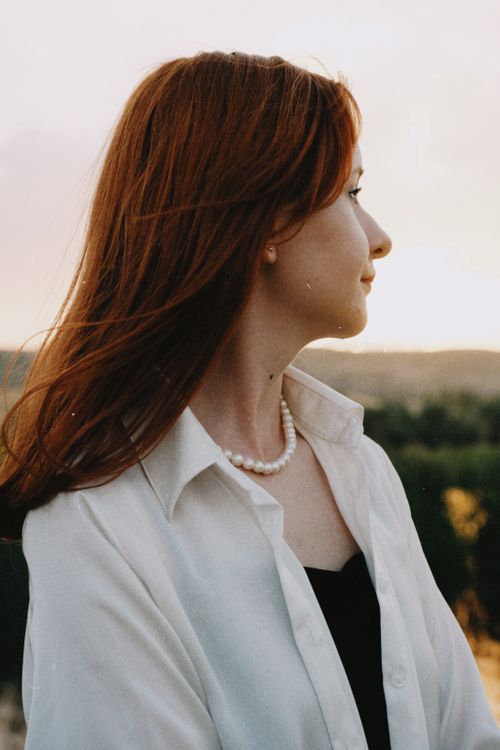 Free Side profile of a woman with red hair standing outdoors, elegantly dressed in a white shirt at sunset. Stock Photo