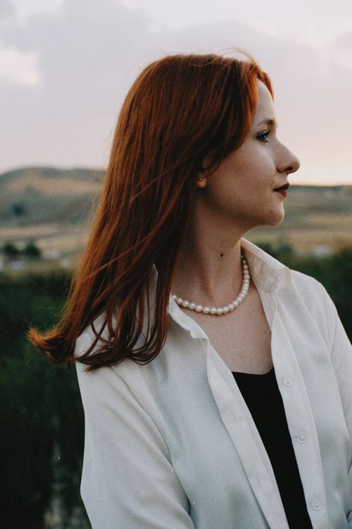 Free Profile portrait of a woman with red hair, wearing a white blouse and pearl necklace, standing outdoors. Stock Photo