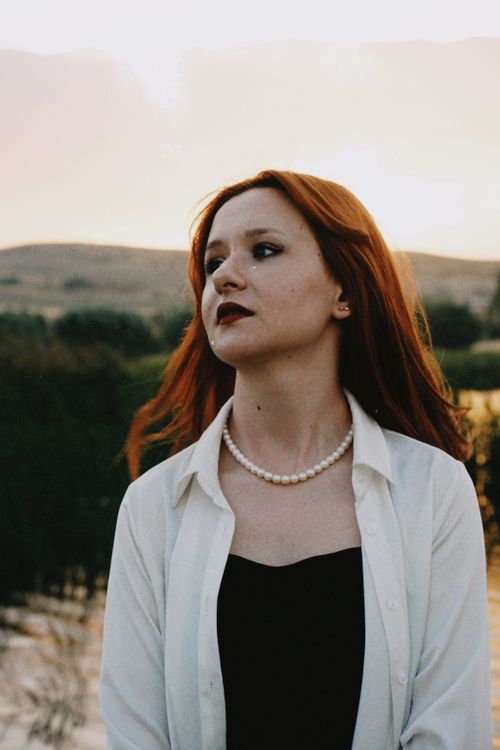 Free Portrait of a redhead woman in a white shirt and pearls, set against an outdoor backdrop at sunset. Stock Photo