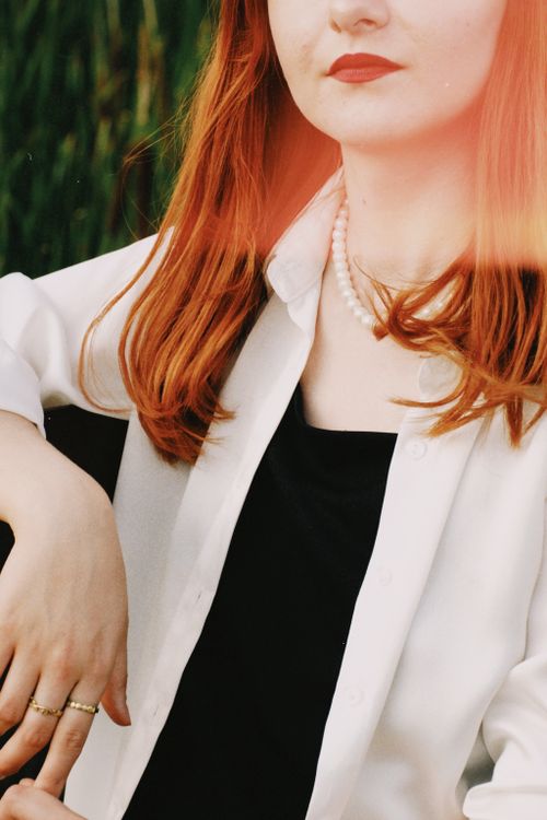 Free A red-haired woman in a white shirt with pearls, posing elegantly outdoors. Stock Photo