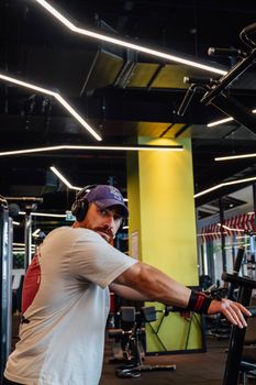 Caucasian man in a gym wearing headphones and a cap, engaged in exercise.