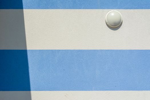 Close-up of a blue and white striped wall with a round wall lamp casting a shadow.