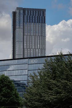 A tall glass skyscraper with modern architecture against a blue sky and clouds, surrounded by trees.