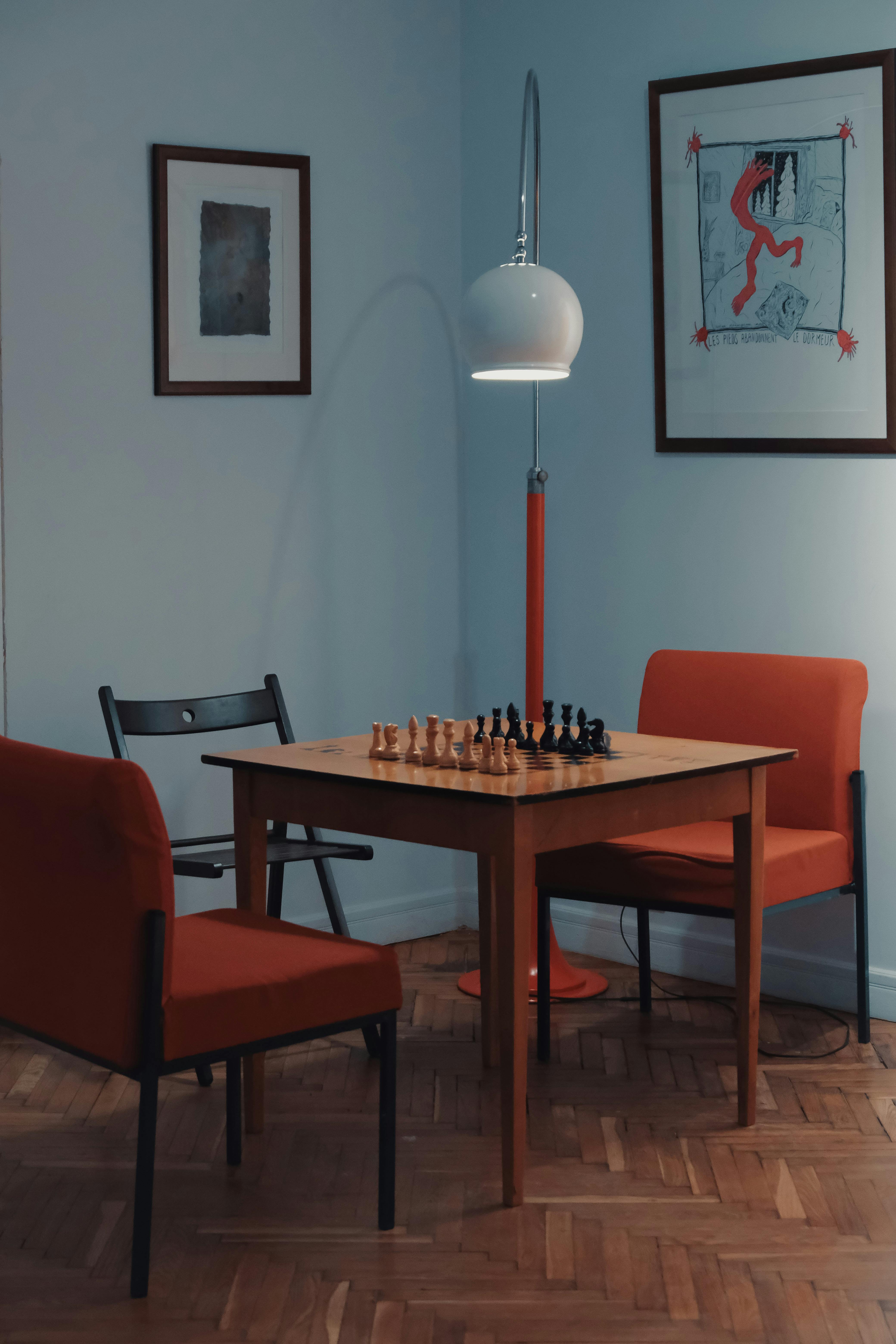 Modern interior with chess set on wooden table, orange chairs, and unique lamp.