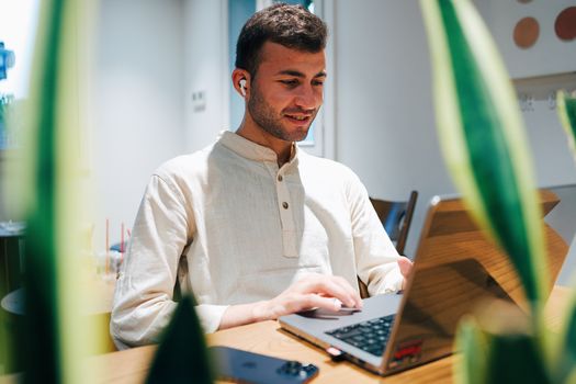 A young man working on a laptop in a modern cafe with wireless earbuds, surrounded by plants.