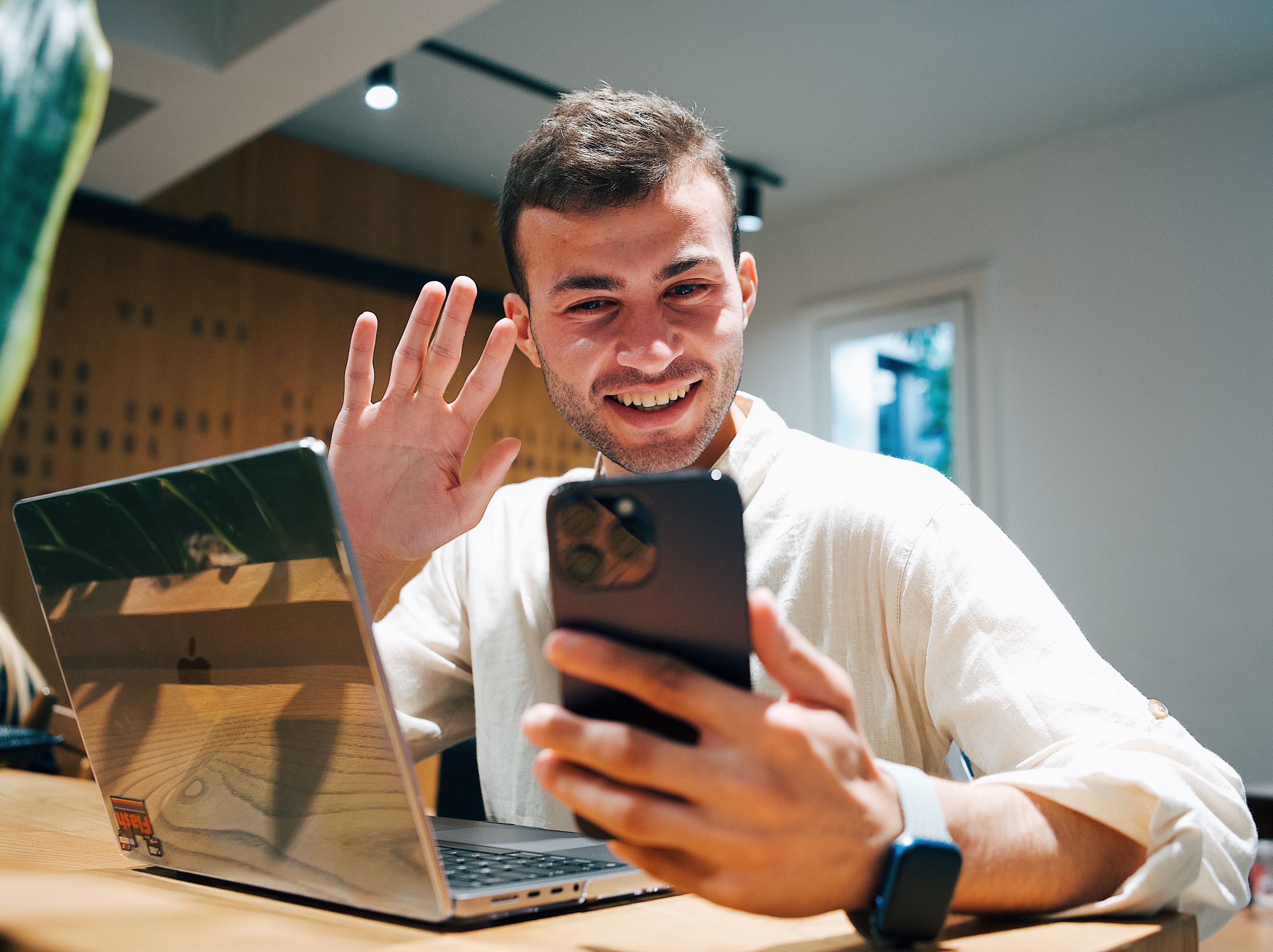 Smiling young man making a video call on his smartphone while sitting with a laptop in a modern office.