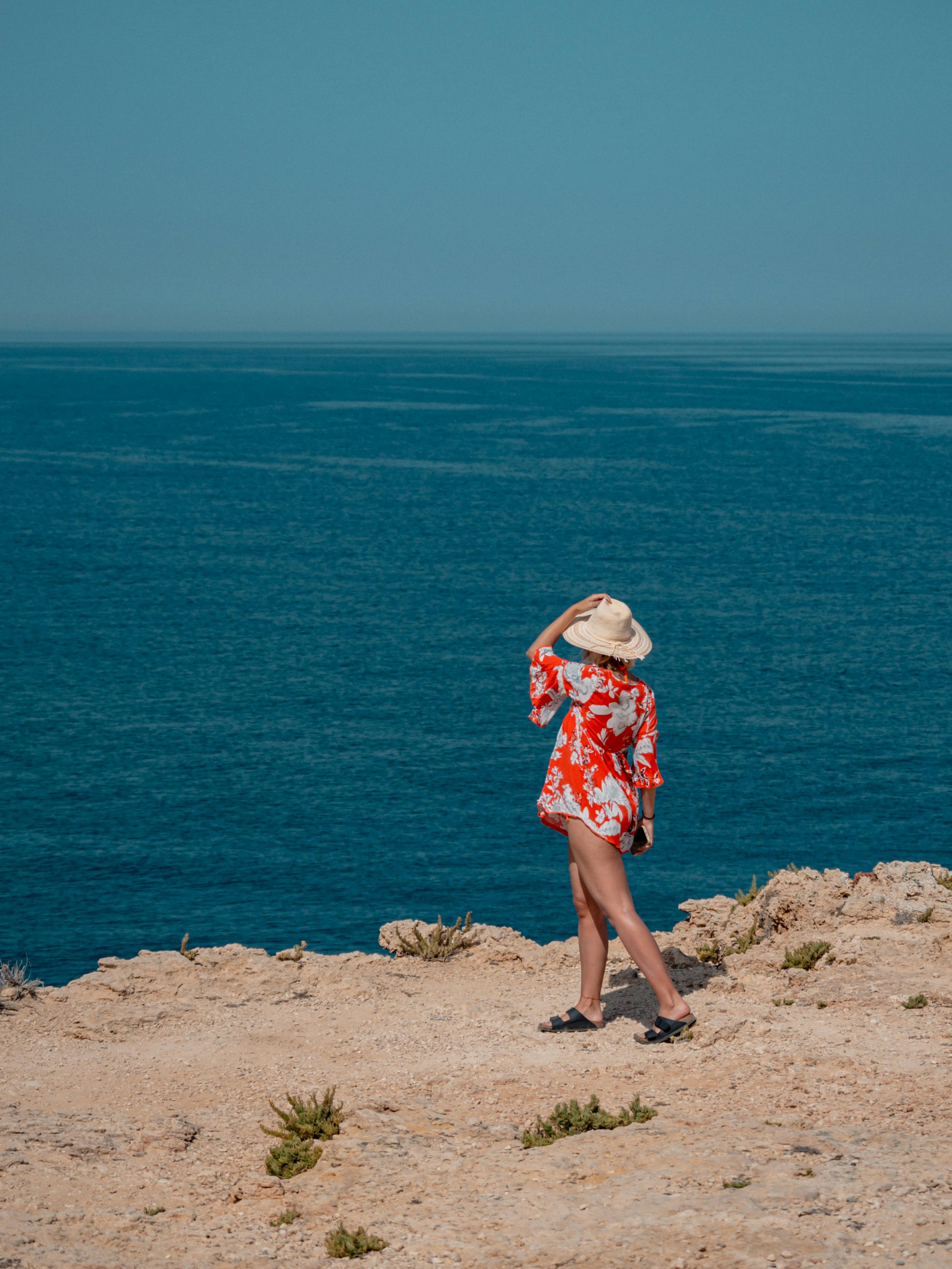 A woman in a red floral dress and hat stands on a rocky cliff by the sea.