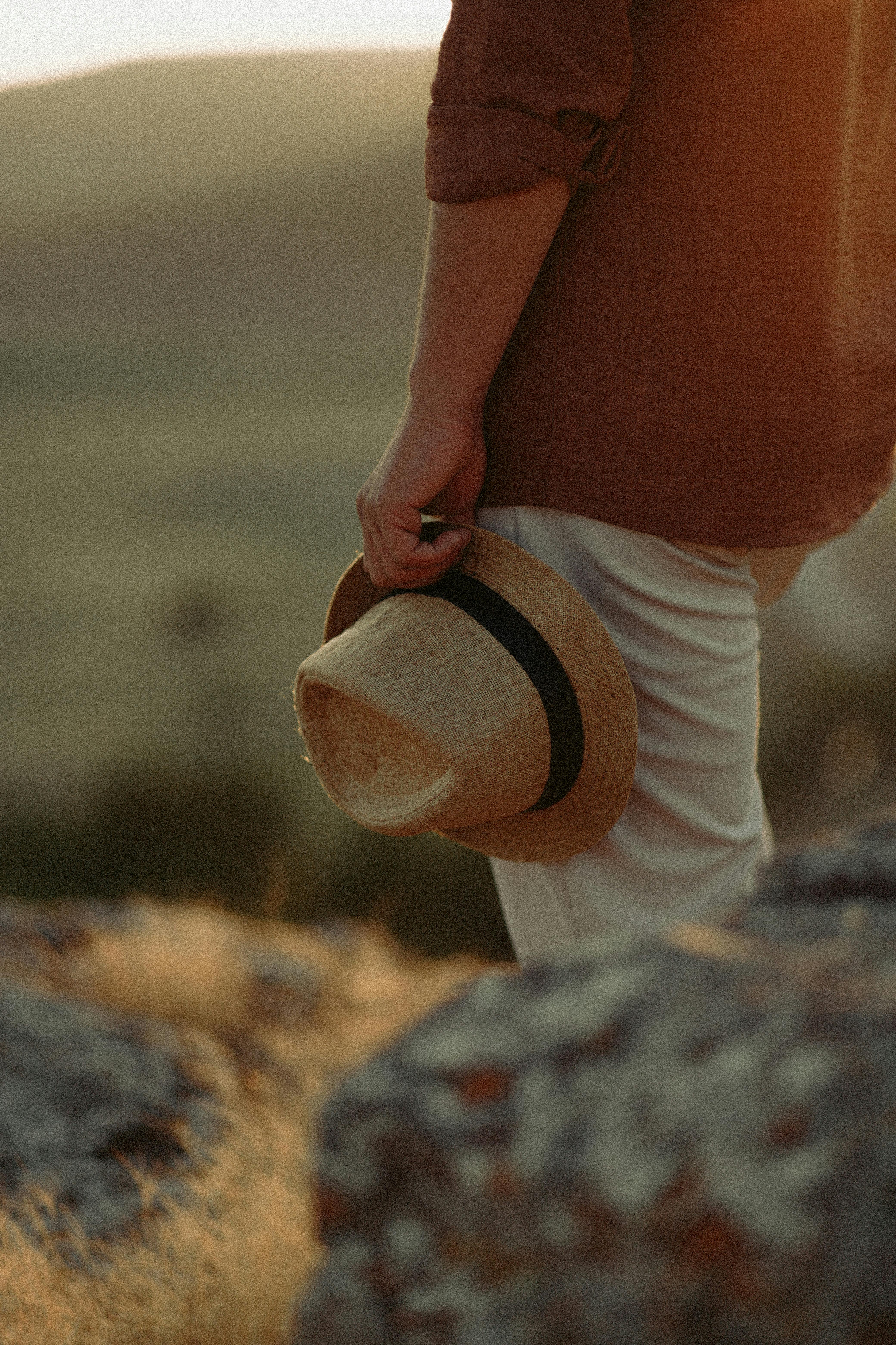 A man stands holding a straw hat in a tranquil outdoor setting during sunset.