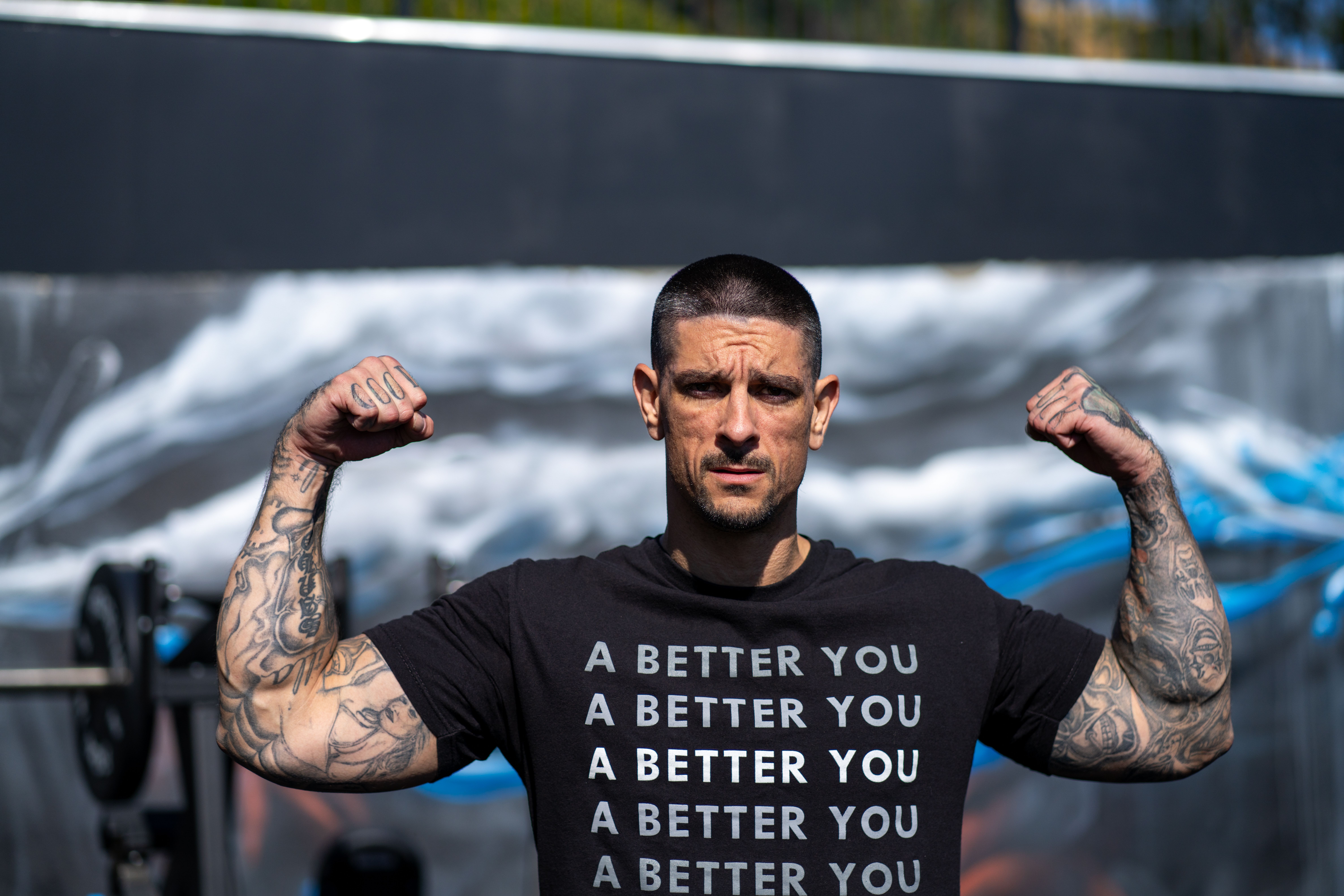 Man flexing muscles outdoors wearing a motivational shirt, tattoos visible, sunny day.