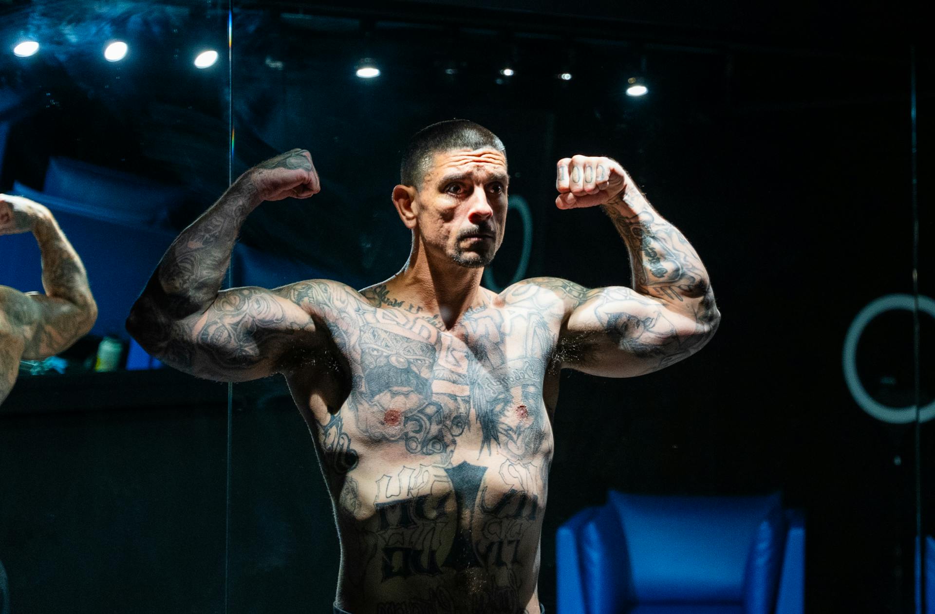 A muscular tattooed man confidently flexing in front of a gym mirror indoors.