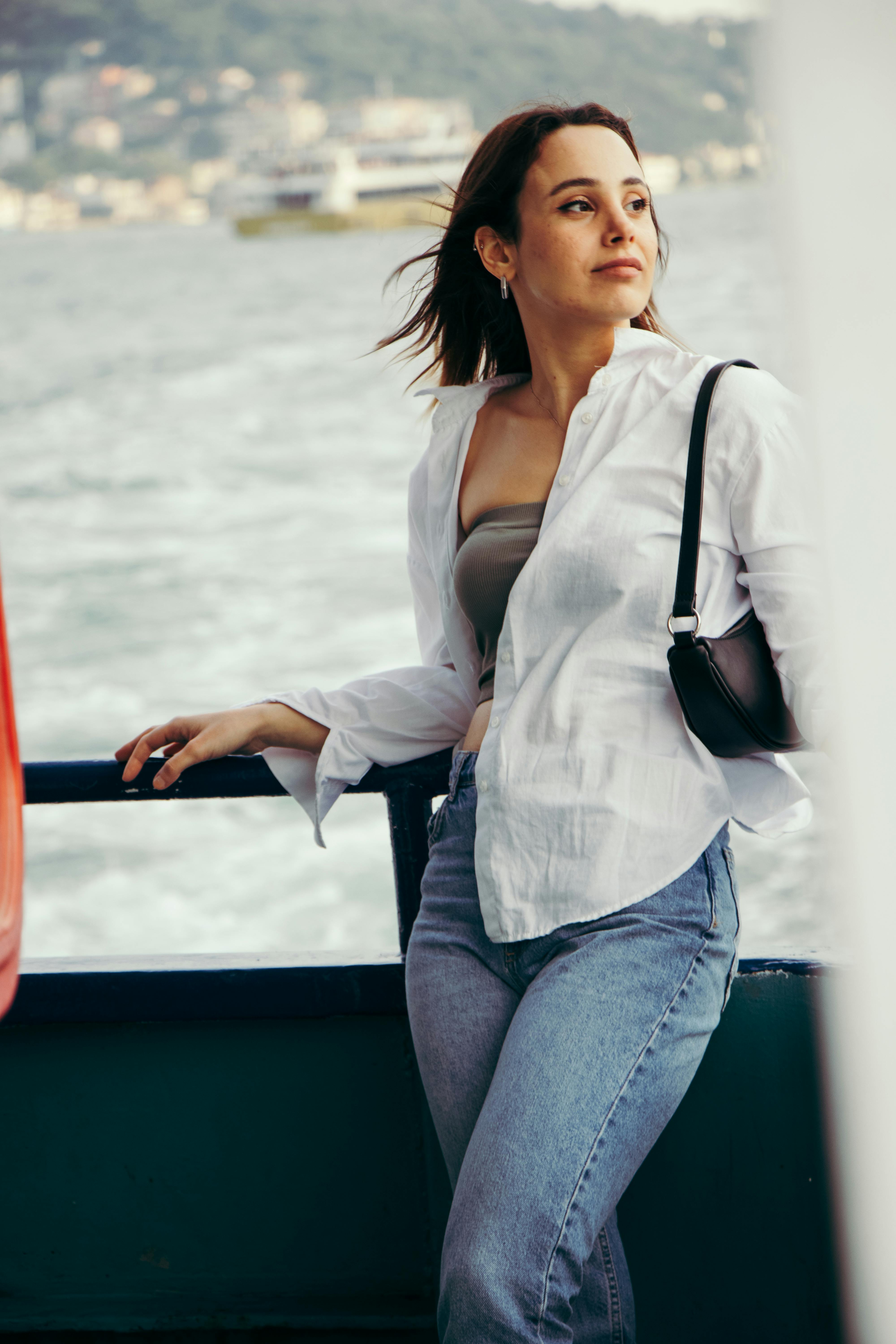 A woman stands on a boat deck, wearing a white shirt and jeans, looking towards the horizon.